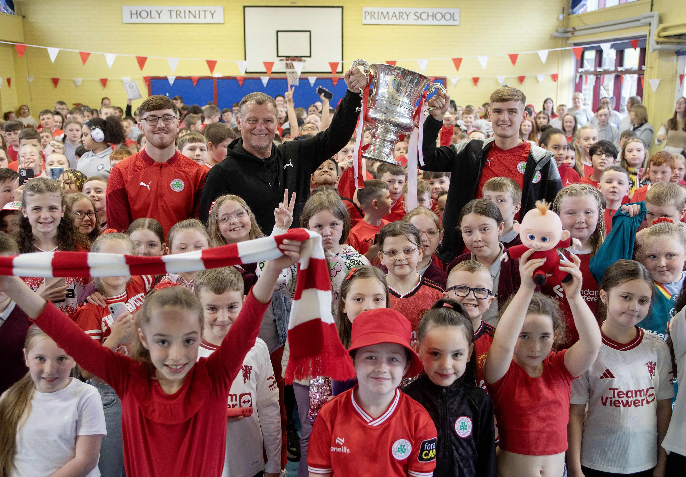 YOU REDS: Cliftonville Manager Jim Magilton with players Odhrán Casey and Sean Stewart and pupils at Holy Trinity PS