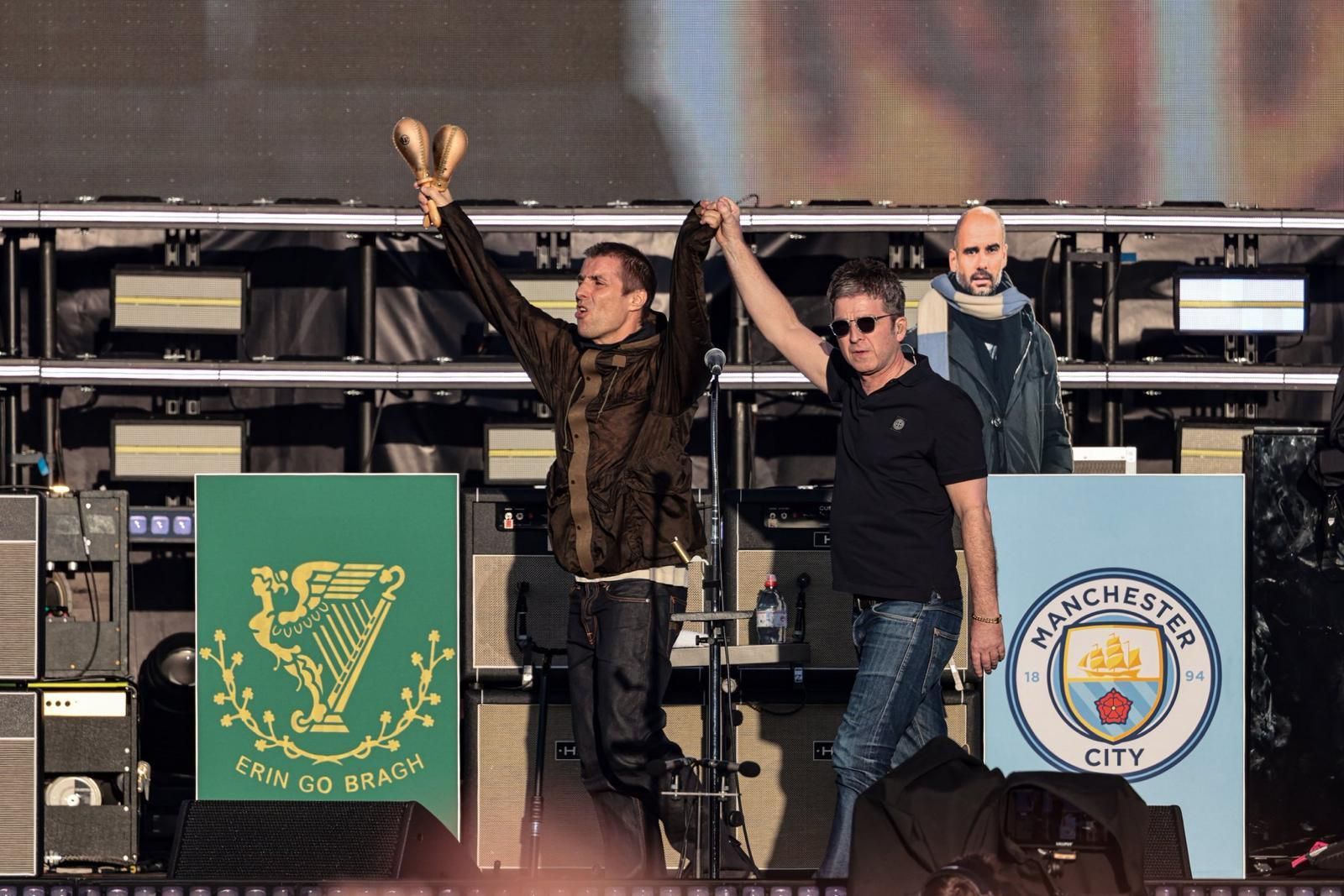 AISÉIRÍ: Noel agus Liam ag teacht ar an ardán, Heaton Park, Manchain agus bratach na nÉireannach Aontaithe sa chúlra.